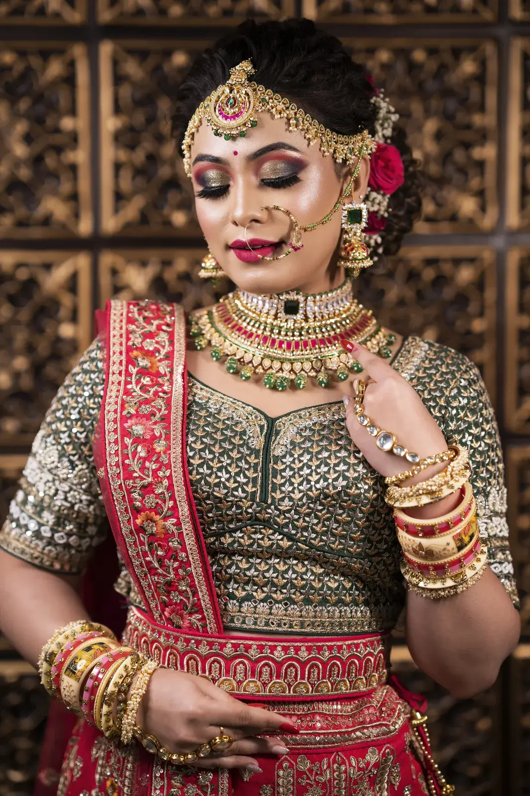 Bride in green and red with maang tikka and nath against a carved wooden screen