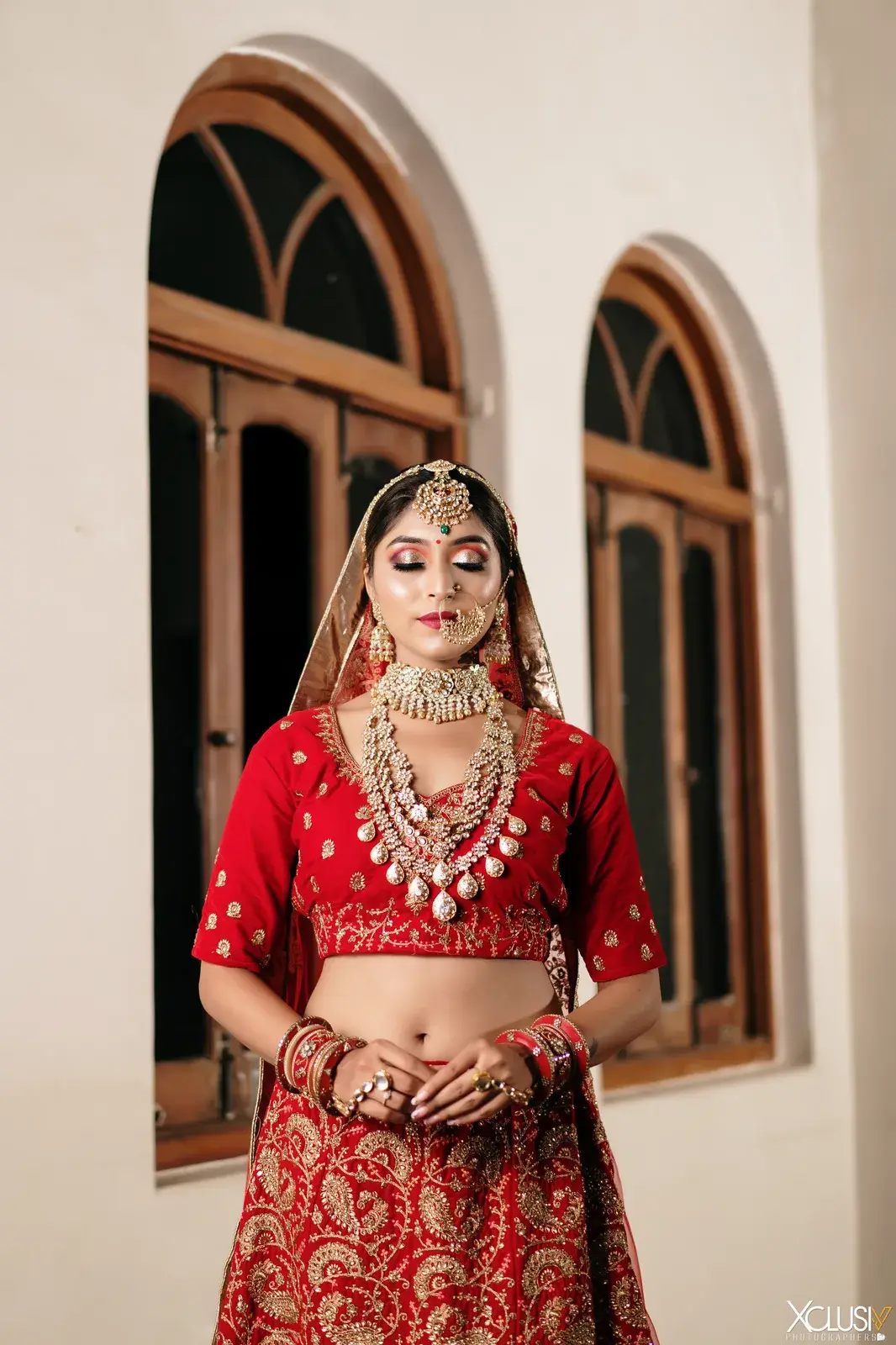 Bride in a red lehenga and layered kundan necklaces, standing by arched windows