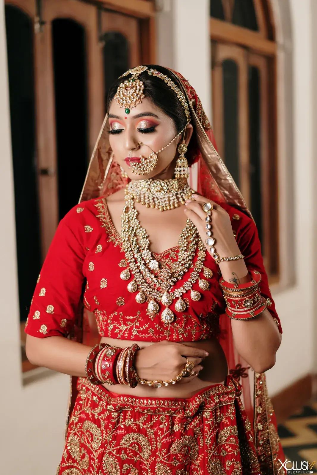 Bride in red with kundan jewellery, looking down, beside arched windows