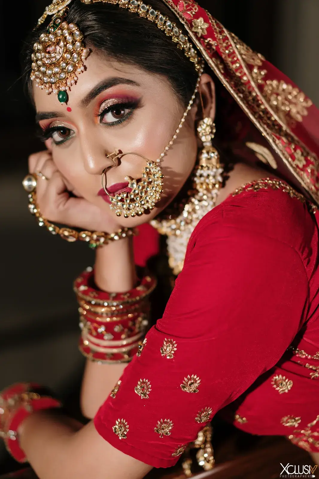 Bride in red resting her chin on her hand, showing red bangles and nath