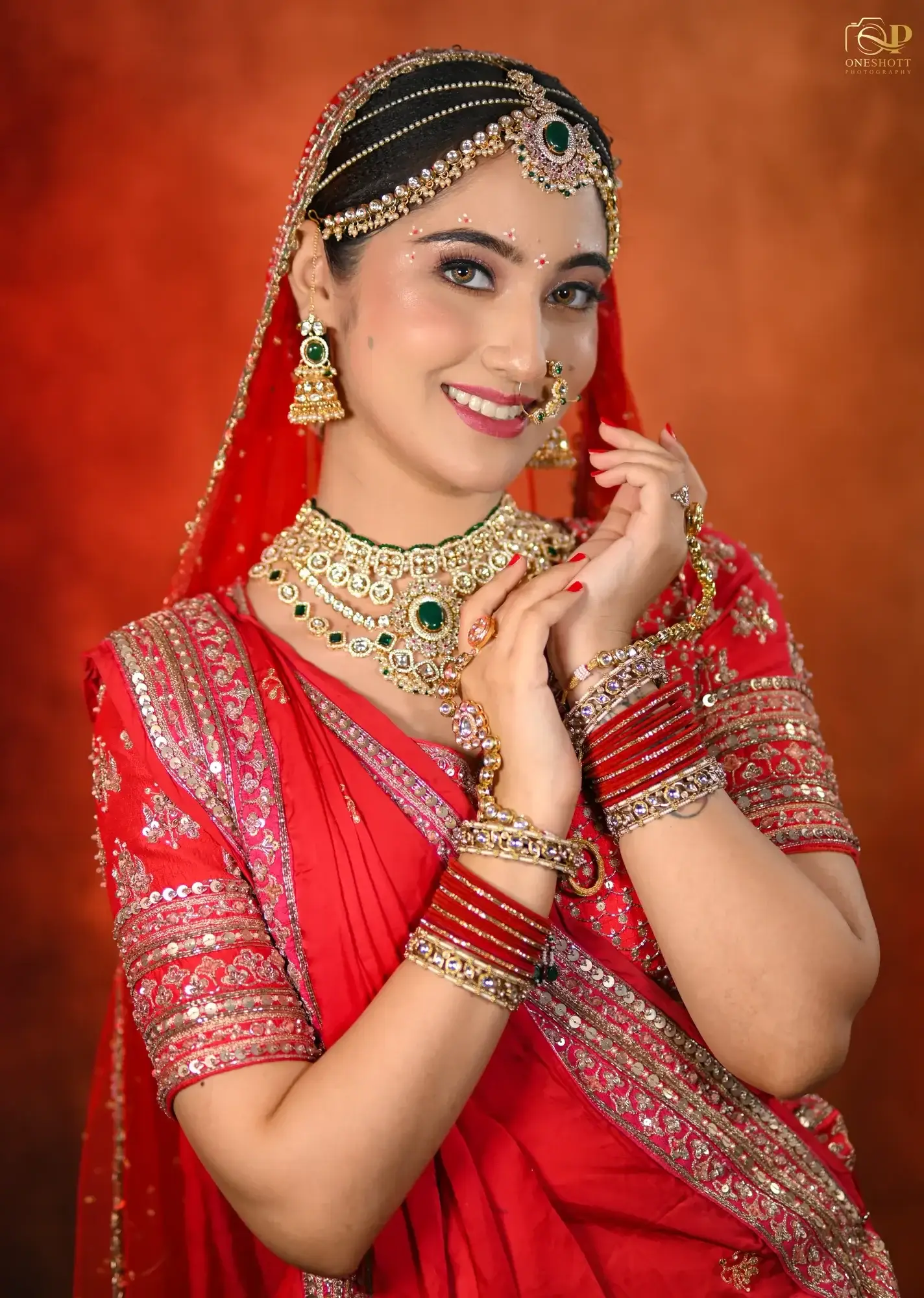 Bride in a red saree with a green-stone necklace and red bangles on a warm red backdrop