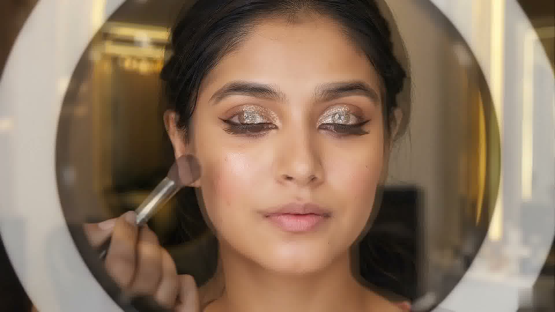 Blush being brushed onto a woman's cheek, her eyes closed under gold glitter eyeshadow